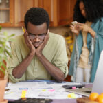 Stressed Young African Male Doing Paperwork At Home, Holding Hands On Head Having Frustrated And Puzzled Look While Reading Notification From Bank In Front Of Him, Failed To Pay Loan In Time