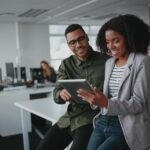 Successful Two African American Young Businesspeople Sitting On Desk Using Digital Tablet While Colleague In Background At Office