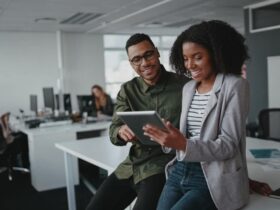 Successful Two African American Young Businesspeople Sitting On Desk Using Digital Tablet While Colleague In Background At Office