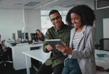 Successful Two African American Young Businesspeople Sitting On Desk Using Digital Tablet While Colleague In Background At Office